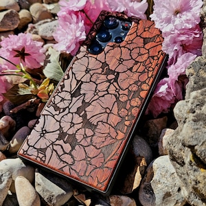 May include: A dark brown wooden phone case with a floral design. The case has a black border and a camera cutout. The case is lying on a bed of small rocks and pink flowers.