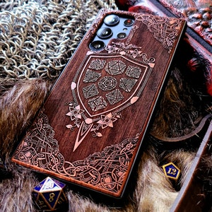 May include: A brown wooden phone case with a detailed design of a shield and dice. The shield has a Celtic knot design and the dice are in the center of the shield. The case is surrounded by a chainmail background.