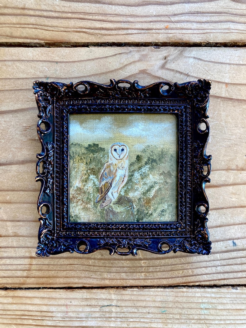 Peut inclure: Une peinture encadr&eacute;e d'un hibou des granges perch&eacute; sur une branche dans un d&eacute;cor bois&eacute;. Le hibou a des plumes blanches avec des marques brunes et des yeux jaunes. La peinture est dans un cadre orn&eacute; brun fonc&eacute;.