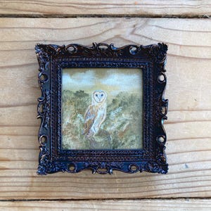Peut inclure: Une peinture encadr&eacute;e repr&eacute;sentant un hibou des granges perch&eacute; sur une branche dans un d&eacute;cor forestier. Le hibou est blanc avec des marques brunes et a des yeux jaunes. La peinture est r&eacute;alis&eacute;e dans un style r&eacute;aliste et se d&eacute;roule sur fond de verdure et de feuillage.