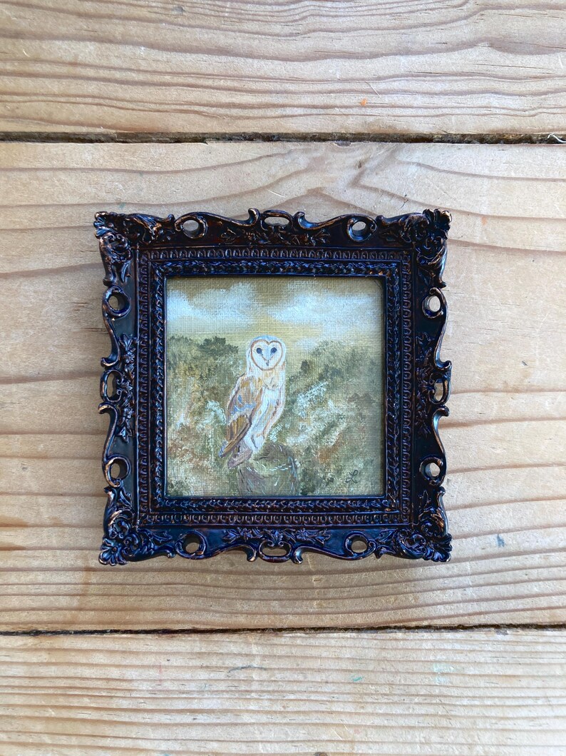 Peut inclure: Une peinture encadr&eacute;e repr&eacute;sentant un hibou des granges perch&eacute; sur une branche dans un d&eacute;cor forestier. Le hibou est blanc avec des marques brunes et a des yeux jaunes. La peinture est r&eacute;alis&eacute;e dans un style r&eacute;aliste et se d&eacute;roule sur fond de verdure et de feuillage.
