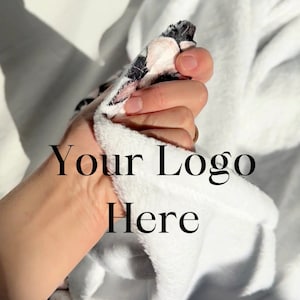 May include: A white plush blanket with a black and pink floral pattern. The blanket is being held by a hand and the text "Your Logo Here" is visible.