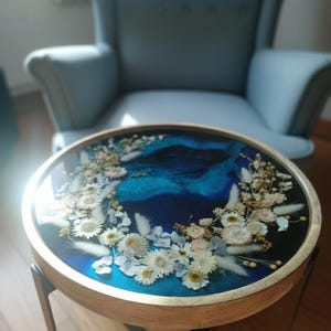 Puede incluir: Mesa auxiliar redonda con marco de madera y tapa de resina azul oscuro. La tapa está decorada con flores secas en blanco, crema y azul claro. La mesa tiene patas de metal negro.