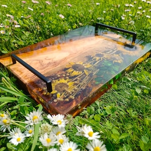 May include: A rectangular serving tray with black handles. The tray has a wooden base with dried sunflowers and other flowers set in clear resin. The tray is on green grass with white and yellow flowers.