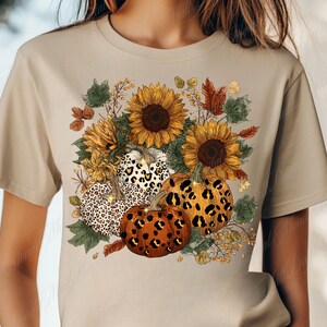 May include: Beige t-shirt featuring a fall-themed graphic. The design includes sunflowers, pumpkins with leopard print patterns, and autumn leaves. The shirt is made of a soft material.