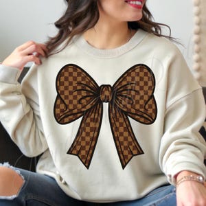 May include: Cream-colored sweatshirt featuring a large brown bow design. The bow has a checkered pattern in shades of brown and is outlined in black. The sweatshirt is a neutral color, making the bow the focal point.