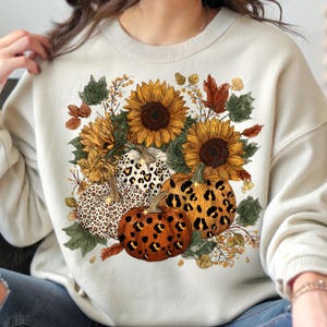 May include: Cream-colored sweatshirt featuring a fall-themed design. The design includes sunflowers, pumpkins with leopard print patterns, and autumn leaves. The pumpkins are orange and white, and the sunflowers are yellow and brown. The overall theme is autumn and harvest.