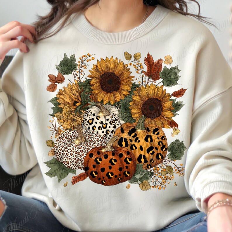 May include: Cream-colored sweatshirt featuring a fall-themed design. The design includes sunflowers, pumpkins with leopard print patterns, and autumn leaves. The pumpkins are orange and white, and the sunflowers are yellow and brown. The overall theme is autumn and harvest.