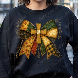 Könnte beinhalten: Schwarzes Sweatshirt mit einem großen Schleifendesign und einem Kreuz in der Mitte. Die Schleife enthält florale, karierte und gemusterte Bänder in Gold-, Grün- und Rottönen. Der Text lautet "God is Within Her, She Will Not Fall" und "Psalm 46:5".