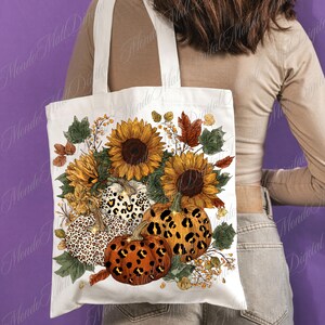 May include: White tote bag featuring a fall-themed design. The design includes sunflowers, pumpkins with leopard print patterns, and autumn leaves. The bag has cream-colored handles and is held by a person.