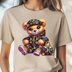 May include: A beige t-shirt with a cartoon illustration of a teddy bear wearing a black and white patterned jacket, a black and white patterned hat, and holding a black and white patterned handbag. The bear is wearing colorful sneakers.