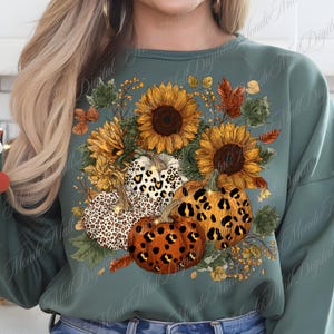 May include: A teal green sweatshirt featuring a fall-themed design. The design includes sunflowers, pumpkins with leopard print patterns, and autumn leaves. The pumpkins are orange, white, and brown. The sunflowers are yellow and brown.