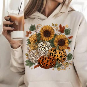 May include: Cream-colored hoodie featuring a fall-themed design. The design includes sunflowers, pumpkins with leopard print patterns, and autumn leaves. The overall aesthetic is warm and seasonal.