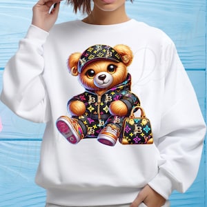 May include: White sweatshirt with a cartoon teddy bear wearing a black and white patterned jacket, a black and white patterned hat, and holding a black and white patterned handbag. The teddy bear has brown fur and is wearing colorful sneakers.