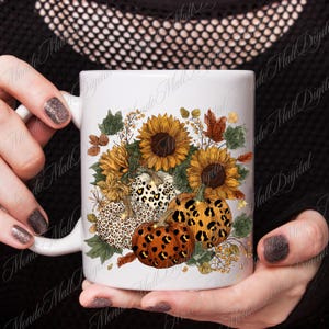 May include: White ceramic mug featuring a fall-themed design. The design includes sunflowers, pumpkins with leopard print patterns, and autumn leaves in shades of orange, brown, and yellow. The mug is held by a person.
