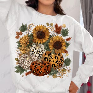 May include: White sweatshirt featuring a fall-themed design. The design includes sunflowers, pumpkins with leopard print patterns, and autumn leaves in shades of orange, brown, and green. The overall theme is a fall harvest.