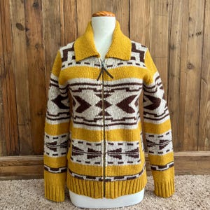 May include: A long-sleeve knit zip-up sweater featuring a mustard yellow, dark brown, and cream geometric pattern. The garment has a fold-over collar, a full-length front zipper with a brown pull tab, and ribbed cuffs and hemline.