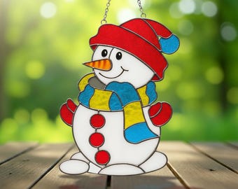 May include: A stained glass snowman ornament with a red hat, scarf, and mittens. The snowman has a carrot nose, black eyes, and a smiling mouth. The ornament is hanging from a silver chain and is set against a blurred green background.
