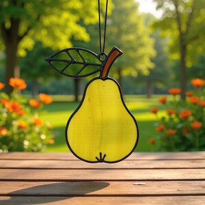 May include: A stained glass pear suncatcher with a black outline, a yellow body, a green leaf, and a brown stem. The suncatcher is hanging in front of a blurred background of trees and flowers.