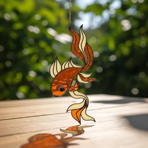 May include: A stained glass goldfish ornament suspended by a chain. The fish has an orange body with cream and orange fins and tail. The ornament is set against a blurred green backdrop, casting a shadow on a wooden surface.