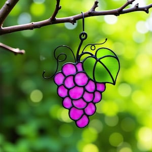 May include: A stained glass grape cluster suncatcher with a pink grape design and a green leaf. The suncatcher is hanging from a branch.