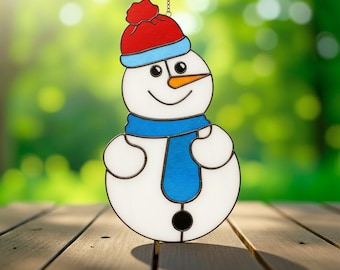 May include: A stained glass snowman ornament with a red hat, blue scarf, and orange carrot nose. The snowman has a smiling face and is hanging from a chain. The background is a blurred green.