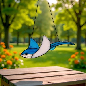 May include: A stained glass manta ray suncatcher, featuring shades of blue, white, and a touch of turquoise. The decorative piece is suspended by black strings, with a detailed design and a smiling face. The background shows a blurred green park.