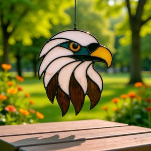 May include: Stained glass eagle head suncatcher with white, brown, yellow, and teal accents. The eagle has a yellow beak and a detailed eye. The suncatcher is hanging from a black string. The background is a blurred outdoor scene.