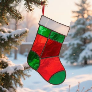 Puede incluir: Un adorno de calcetín navideño de vidriera en rojo, verde y blanco cuelga de una cinta roja. El adorno está suspendido frente a una escena invernal nevada, con un fondo borroso de árboles y una casa.