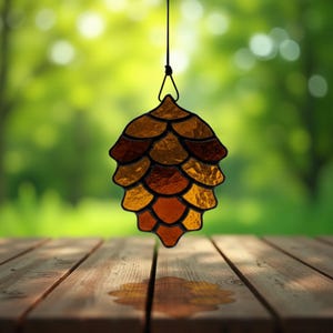 May include: A stained glass pinecone ornament in shades of amber, brown, and gold hangs from a black string. The ornament is suspended against a blurred green background, with a wooden table in the foreground.