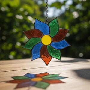 May include: A stained glass flower suncatcher with a yellow centre and petals in blue, green, orange, and brown. The decorative piece hangs from a black chain. The suncatcher's reflection is visible below.