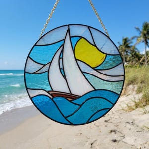May include: Round stained glass art piece depicting a sailboat on the ocean with a yellow sun. The artwork features a white sailboat with a brown hull, set against blue and turquoise water and sky. A gold chain is attached for hanging.