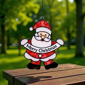 May include: A Christmas decoration in the shape of Santa Claus, with a red hat and suit, white beard, and the words "Merry Christmas" on a banner. The ornament is hanging against a blurred green backdrop.