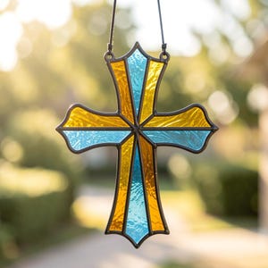 Pode incluir: Um ornamento de cruz em vitral com painéis alternados de vidro texturizado em amarelo âmbar e azul claro. As peças são unidas por solda de metal escuro, formando uma cruz decorativa suspensa por um cordão preto fino para pendurar.