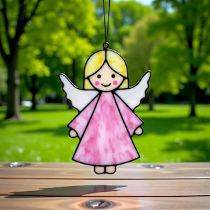 Könnte beinhalten: Ein Glasengel-Ornament mit schwarzer Umrandung. Der Engel hat einen gelben Kopf, weiße Flügel und ein rosa Kleid. Das Ornament hängt vor einem verschwommenen grünen Hintergrund.