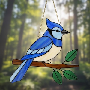 以下が含まれることがあります： 金色のチェーンから吊り下げられたステンドグラスの青い鳥のサンキャッチャー。鳥は緑の葉が付いた茶色の枝に止まっています。鳥は黒い嘴と目をしており、白と水色のアクセントがあります。