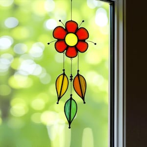 May include: A stained glass suncatcher with a red flower and three leaves in shades of green, yellow, and orange. The suncatcher is hanging from a window.