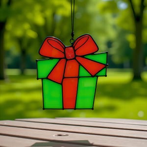 May include: A stained glass ornament in the shape of a gift box with a red bow. The box is green, and the bow is red. The ornament is hanging from a black string against a blurred green background.