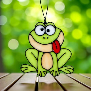May include: A decorative green frog ornament with a red tongue, black outlines, and large white eyes. The frog is suspended from a black string. The background is a blurred green, suggesting an outdoor setting. The frog's design is similar to stained glass.