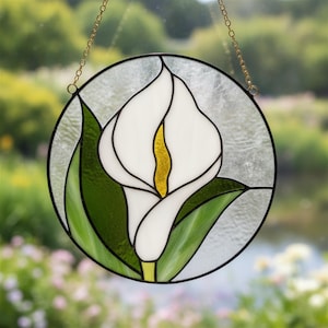 May include: A circular stained glass suncatcher featuring a white calla lily with a yellow spadix and green leaves. The piece is framed in black metal and suspended by a gold-toned metal chain, showcasing textured clear glass in the background.