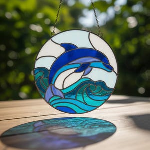 May include: A circular stained glass panel depicting a blue dolphin leaping over turquoise waves. The dolphin is set against a white and light blue background. The panel is suspended by a chain, creating a decorative ocean-themed piece.