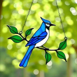 May include: A stained glass blue jay bird suncatcher. The bird is perched on a branch with green leaves. The bird has a white chest and a blue crest. The suncatcher hangs from a black chain.