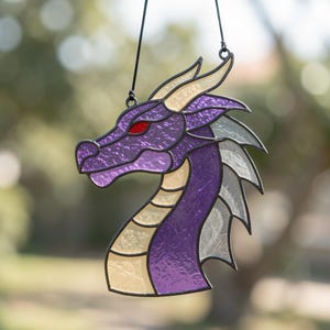 May include: A stained glass suncatcher shaped like a dragon head in profile. The dragon features textured purple glass for the face and neck, cream-colored glass for the underbelly and horns, and translucent white glass for the fins, with a red glass eye.