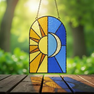 Op de afbeelding: Gebrandschilderd glazen hangpaneel met zon en maan. Het paneel is verticaal verdeeld, met een gele en oranje zon aan de linkerkant en een blauwe en gouden maan aan de rechterkant. Het paneel is omlijst met donker metaal en hangt aan een ketting.