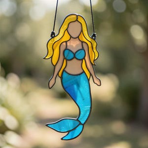 Peut inclure: Un attrape-soleil en vitrail en forme de sirène avec de longs cheveux jaunes, un haut de bikini bleu et une queue bleue. La silhouette est soulignée par une soudure métallique sombre et suspendue par un cordon noir, avec des panneaux en verre translucide.
