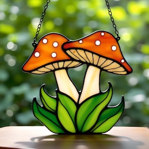 Puede incluir: Atrapasoles de vidrio manchado con dos setas. Las setas tienen sombreros naranjas con puntos blancos y tallos color crema. El diseño se completa con hojas verdes y cuelga de una cadena negra. Decoración para ventanas o paredes.