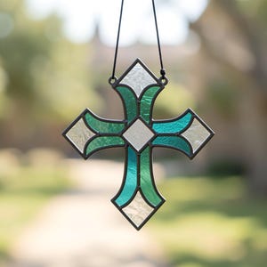 Pode incluir: Um ornamento de cruz em vitral com uma forma de diamante central rodeada por quatro braços curvos. O design incorpora painéis de vidro branco translúcido e secções de vidro verde-azulado vibrante, unidos por linhas de soldadura de metal escuro.