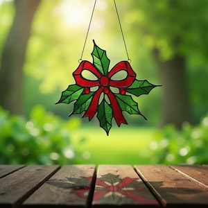 May include: A stained glass Christmas ornament featuring a red bow and green holly leaves. The ornament is suspended by a black string against a blurred green background. The bow has red accents, and the leaves are green.