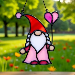 May include: A stained glass gnome with a red hat, white beard, and pink body, holding a pink heart-shaped balloon. The gnome is outlined in black and has a black hanging string. The background is a blurred green field with flowers.
