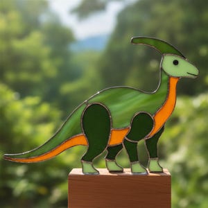 Peut inclure: Une figurine de dinosaure parasaurolophus en vitrail composée de nuances de vert clair et foncé avec des accents orange le long du cou, du ventre et de la queue. Le dinosaure est monté sur une base rectangulaire en bois avec des soudures métalliques.